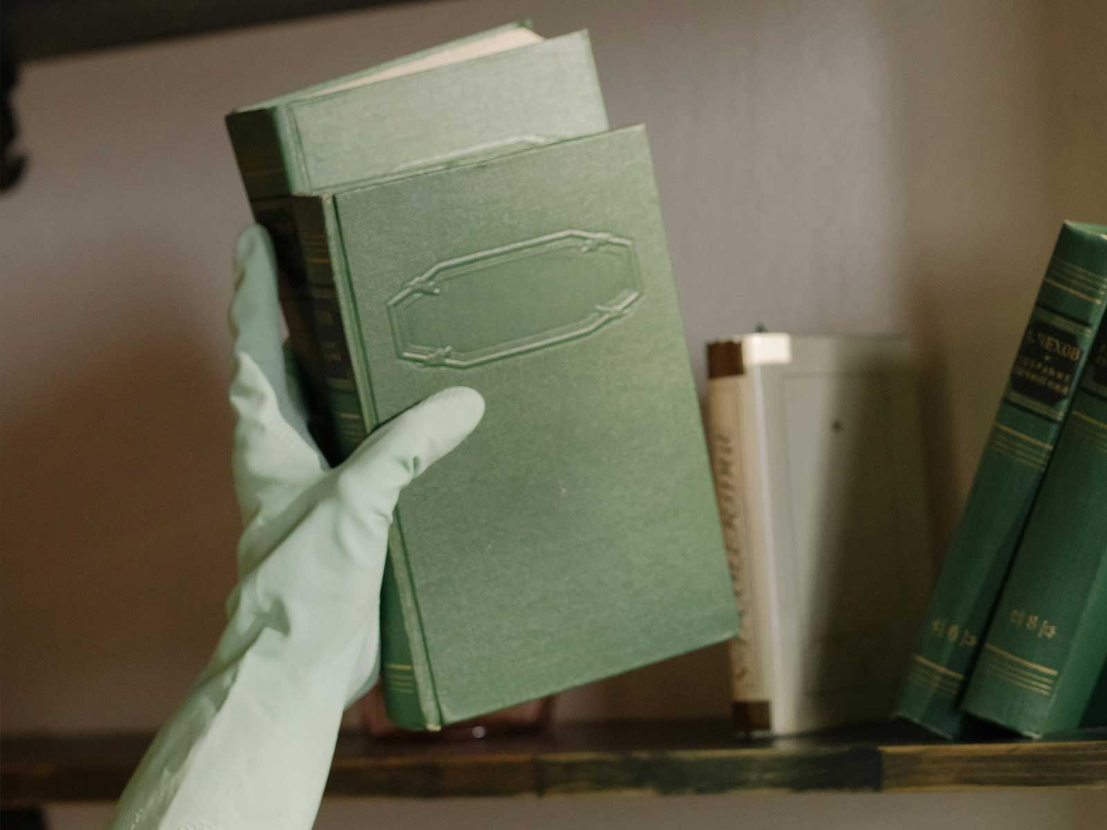 A gloved hand carefully lifting a green book from a shelf, symbolizing mindful dusting and thoughtful home upkeep with Lenuma.