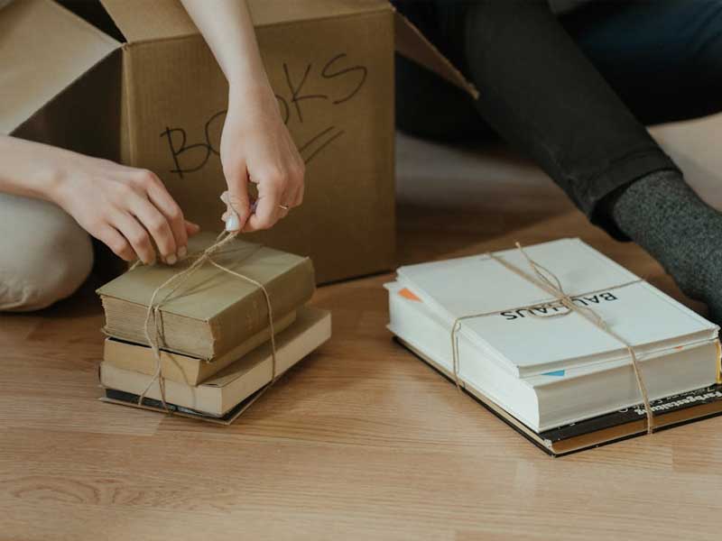 Hands carefully bundling books with twine beside a labeled moving box, illustrating mindful, respectful home care during moving cleaning.
